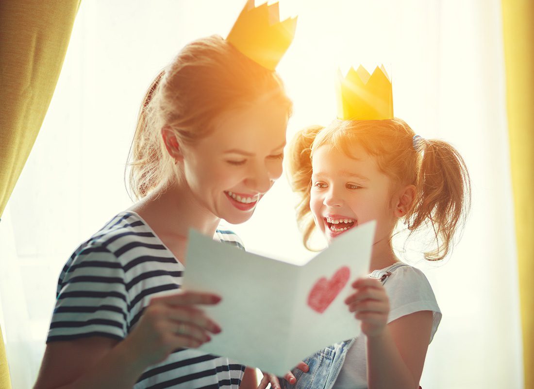 Read Our Reviews - Portrait of a Cheerful Mother Reading a Hand Drawn Card with a Heart on it From Her Happy Daughter Both Wearing Crowns at Home