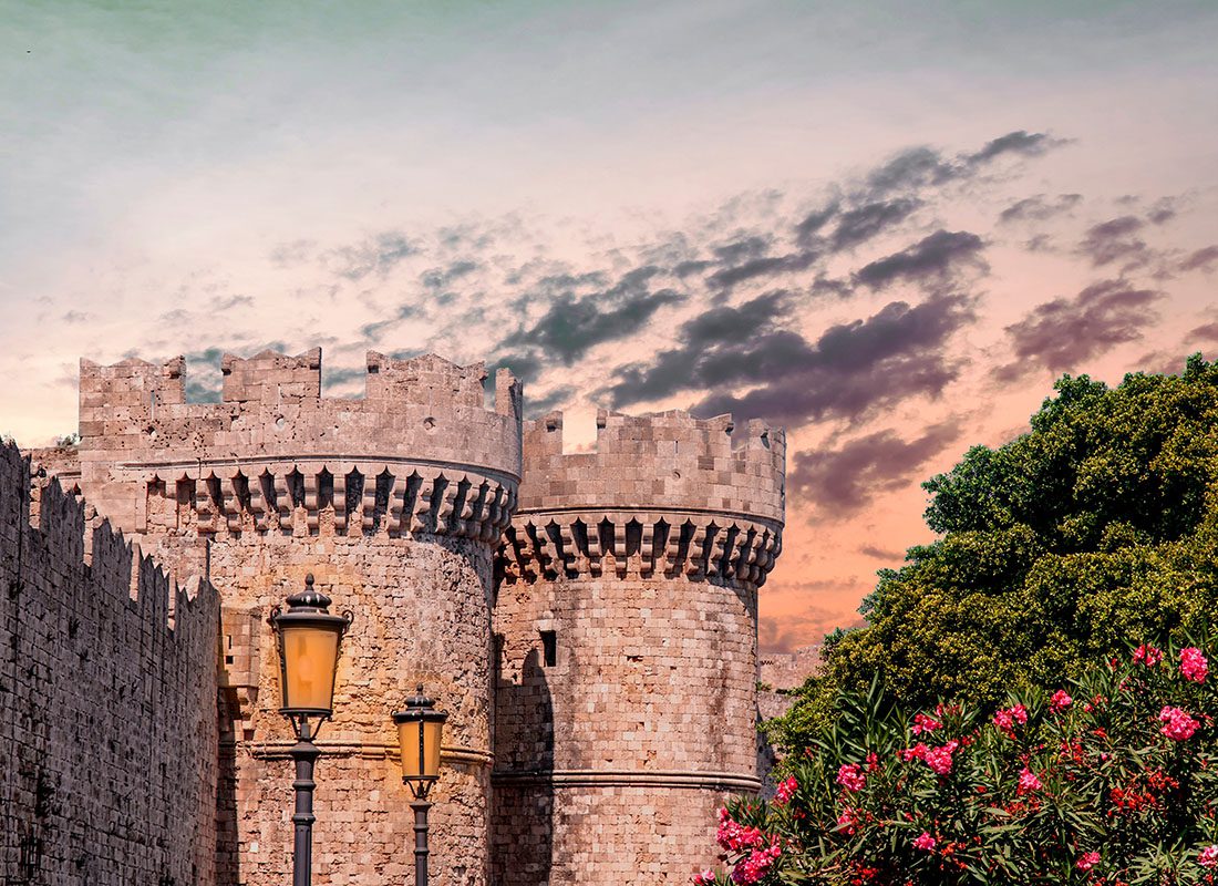 We Are Independent - Scenic View of a Castle with Two Towers Against a Colorful Sunset Sky with Lamp Posts in Front and Flowers